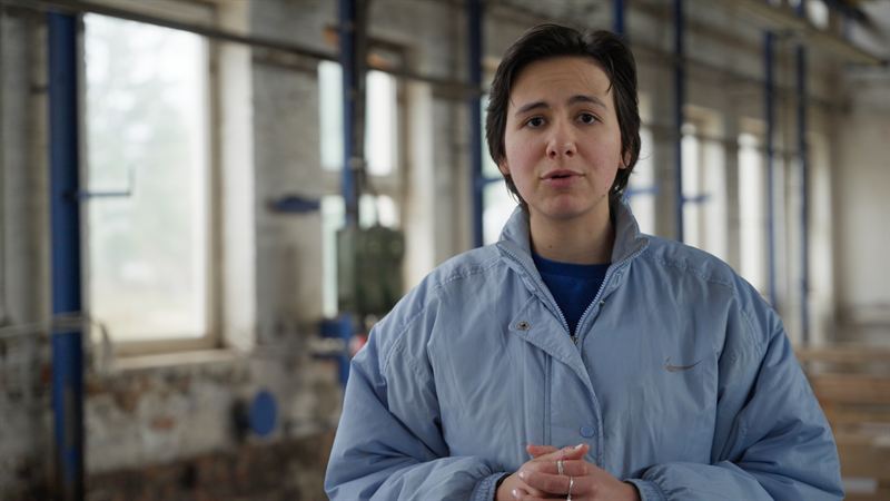 Eine junge Frau in hellblauer Jacke spricht direkt in die Kamera in einer verlassenen Industriehalle – Szene aus der EB-Produktion 'Funk – Der Dunkle Parabelritter'