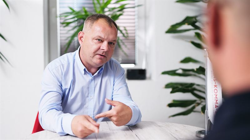 Ein Mann im hellblauen Hemd sitzt gestikulierend an einem Tisch im Gespräch, im Hintergrund Grünpflanzen und ein Banner mit dem Schriftzug des Auftraggebers – Interviewszene aus der Imagefilmproduktion für 1komma1