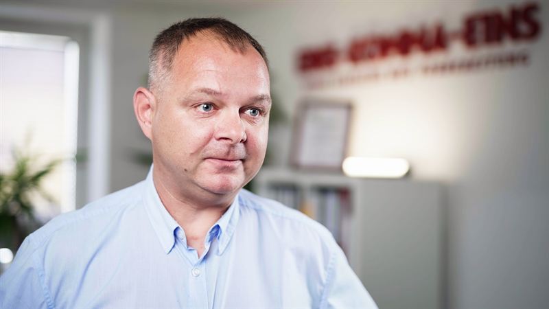Porträt eines Mannes im hellblauen Hemd, im unscharfen Hintergrund ein Banner mit dem Schriftzug des Auftraggebers und eine gerahmte Urkunde – Szene aus der Imagefilmproduktion für 1komma1