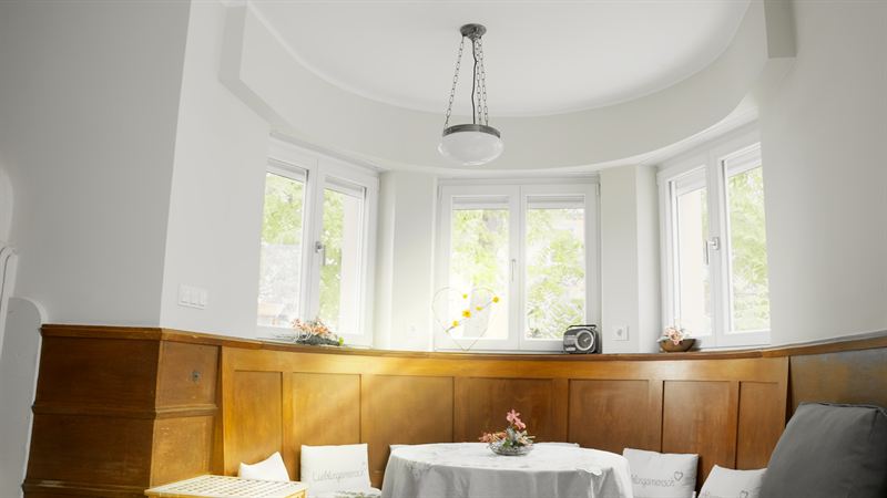 Heller Wohnbereich mit Erkerfenstern, holzverkleideten Wänden und liebevoll gedecktem Tisch mit Blumendeko – Szene aus der Imagefilmproduktion für Pflegedienst Silvia Haake