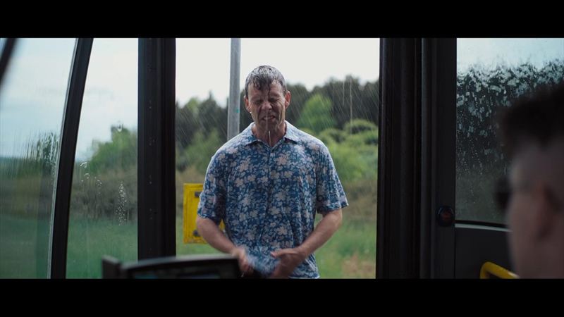 Ein Mann in durchnässtem Blumenhemd steht im Regen vor einer geöffneten Bustür, im Hintergrund grüne Landschaft – Filmszene aus der Imagefilmproduktion für PRG Greiz