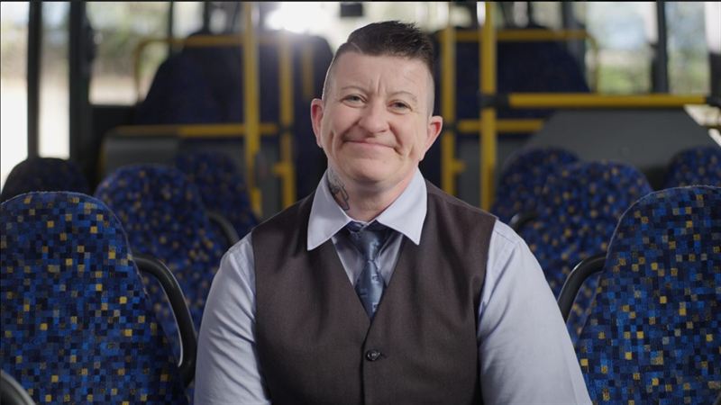 Ein lächelnder Mann mit Weste und Krawatte sitzt in einem Reisebus mit blau gemusterten Sitzen – Interviewaufnahme aus der Imagefilmproduktion für PRG Greiz