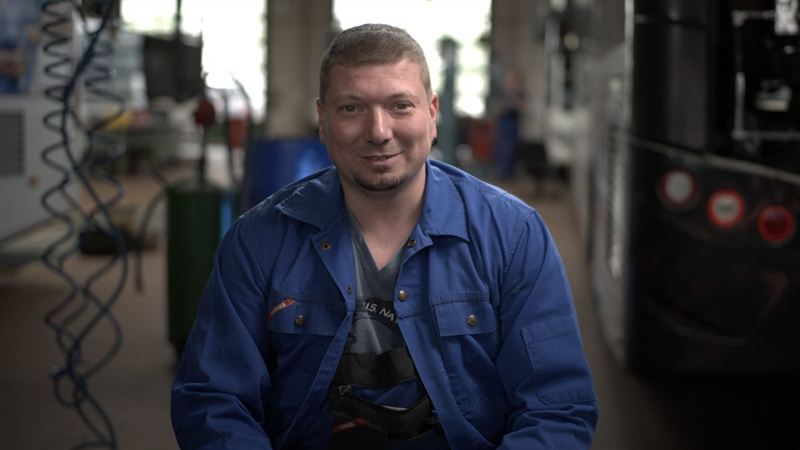 Ein lächelnder Mann in blauer Arbeitsjacke steht in einer Werkstatt – Interviewaufnahme aus der Imagefilmproduktion für PRG Greiz