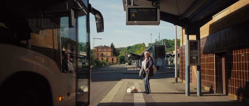 Eine ältere Frau mit Gehstock wartet an einer Bushaltestelle, im Vordergrund spiegelt sich ein Bus – Filmszene aus der Imagefilmproduktion für PRG Greiz
