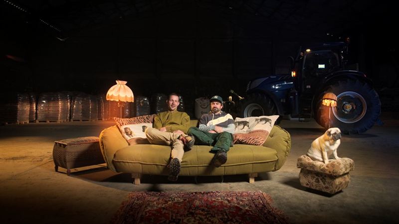 Zwei Männer sitzen auf einem grünen Sofa neben einem Traktor in einer dunklen Halle, daneben ein Mops auf einem Hocker – inszenierte Szene aus der Imagefilmproduktion für Pulsschlag Erde e.V.