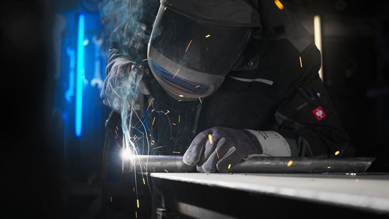 Ein Schweißer mit Schutzmaske arbeitet an einem Metallträger, Funken sprühen im dunklen Werkstattlicht – Testshooting-Aufnahme für Modbord