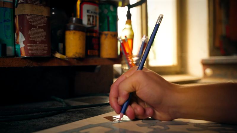 Nahaufnahme einer Hand, die mit einem Bleistift auf Papier zeichnet, im unscharfen Hintergrund Werkstattmaterial – Szene aus dem Kurzfilm 'Der Schwibbogen – Die Tradition lebt'