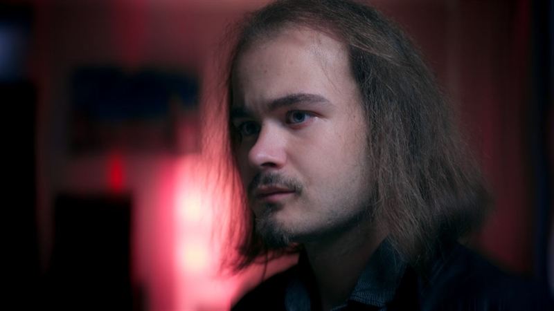 Nahaufnahme eines jungen Mannes mit langen Haaren in rot-blauem Kinolicht, ernster Blick zur Seite – Filmstill aus dem Kurzfilm 'Into Dust – Begraben'