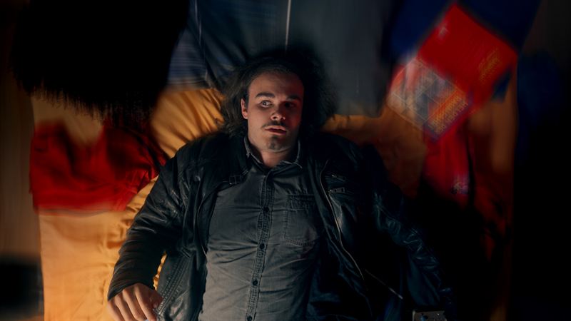 Nahaufnahme eines Mannes mit wilder Frisur und schwarzer Lederjacke vor bunten Fahnen/Tüchern, direkter, eindringlicher Blick in die Kamera – Filmstill aus dem Kurzfilm 'Into Dust – Begraben'