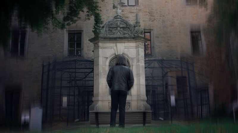 Eine Person geht von hinten auf ein steinernes Brunnendenkmal vor einer historischen Fassade zu, unscharfe Zweige im Vordergrund – Filmstill aus dem Kurzfilm 'Into Dust – Begraben'