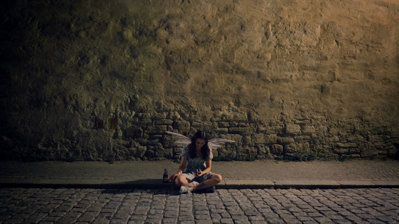 Eine Frau mit flügelartigem Kostüm sitzt nachts auf dem Kopfsteinpflaster vor einer alten Stadtmauer – Filmstill aus dem Kurzfilm 'Into Dust – Begraben'