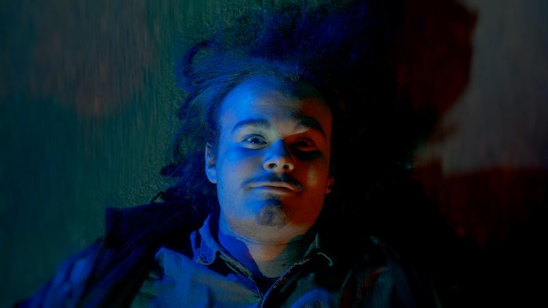 Nahaufnahme eines liegenden Mannes mit wilder Frisur in blau-rotem Kinolicht, starrer Blick nach oben – Filmstill aus dem Kurzfilm 'Into Dust – Begraben'