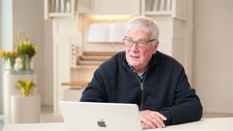Ein älterer Mann mit Brille sitzt vor einem Laptop, im unscharfen Hintergrund eine Kirchenorgel – Weiterbildungsvideo für die Neuapostolische Kirche