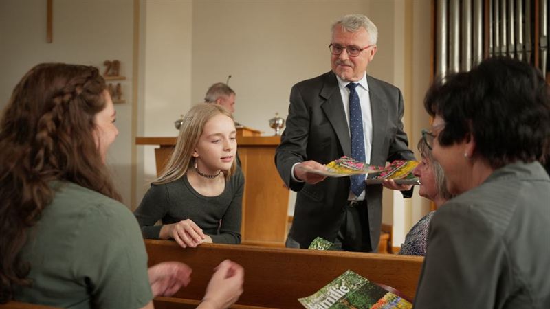 Ein Mann im Anzug übergibt Kindermagazine an eine Gruppe in Kirchenbänken – Szene aus dem Werbefilm für den Verlag Friedrich Bischoff